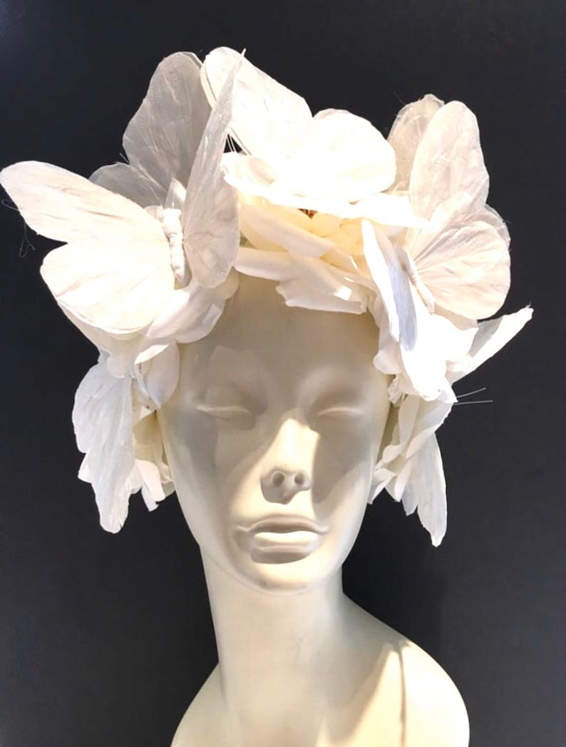 May include: A white headpiece made of fabric flowers and butterflies. The headpiece is worn on a white mannequin head.
