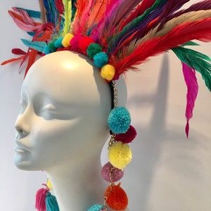 May include: A colorful feather headdress with a headband and dangling pom-poms. The headdress is made of feathers in various shades of red, orange, yellow, green, blue, and purple. The pom-poms are in shades of yellow, orange, pink, red, and blue.