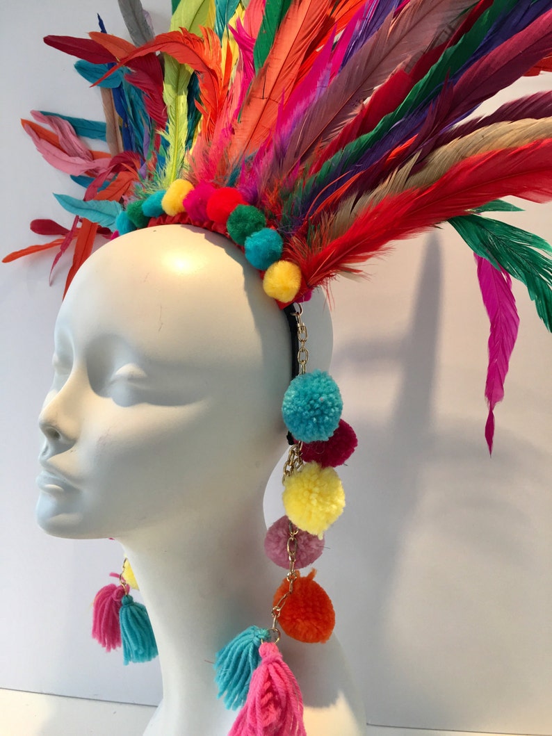 May include: A colorful feather headdress with a headband and dangling pom-poms. The headdress is made of feathers in various shades of red, orange, yellow, green, blue, and purple. The pom-poms are in shades of yellow, orange, pink, red, and blue.