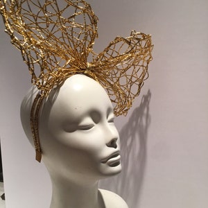 May include: A gold wire mesh headband with a large bow on top. The headband is on a white mannequin head.