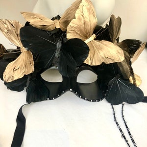 Puede incluir: Una m&aacute;scara negra de carnaval adornada con mariposas doradas y negras. La m&aacute;scara presenta un dise&ntilde;o curvo con peque&ntilde;os detalles brillantes en los bordes y cintas negras para sujetarla.
