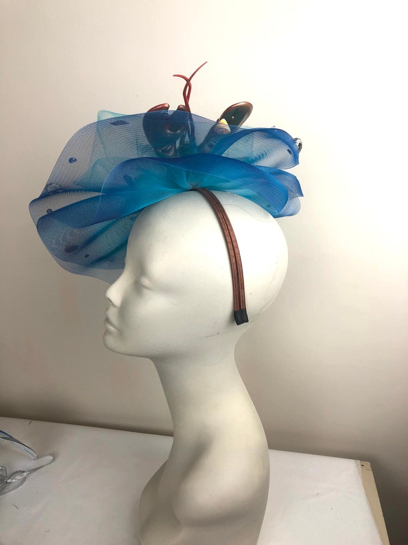 Op de afbeelding: Een blauwe tule hoofdband met een kreeftdecoratie. De hoofdband wordt gedragen op een wit mannequinhoofd.