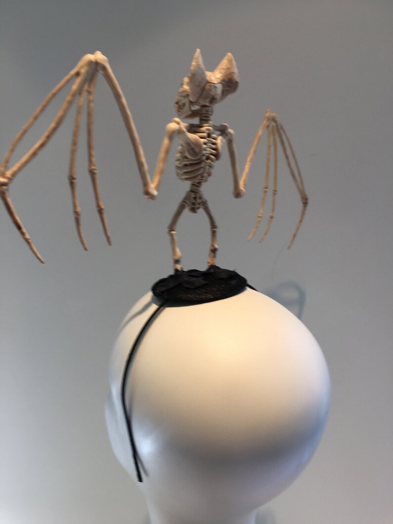 May include: A Halloween-themed headpiece featuring a skeletal bat perched atop a white, rounded form. The bat has outstretched wings and a detailed bone structure. A black, circular accent sits beneath the bat, with a thin black band extending down the sides.