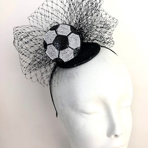 May include: A black headband with a decorative soccer ball design. The soccer ball is embellished with sparkling white and black rhinestones. A black netting is attached to the headband, creating a dramatic effect.