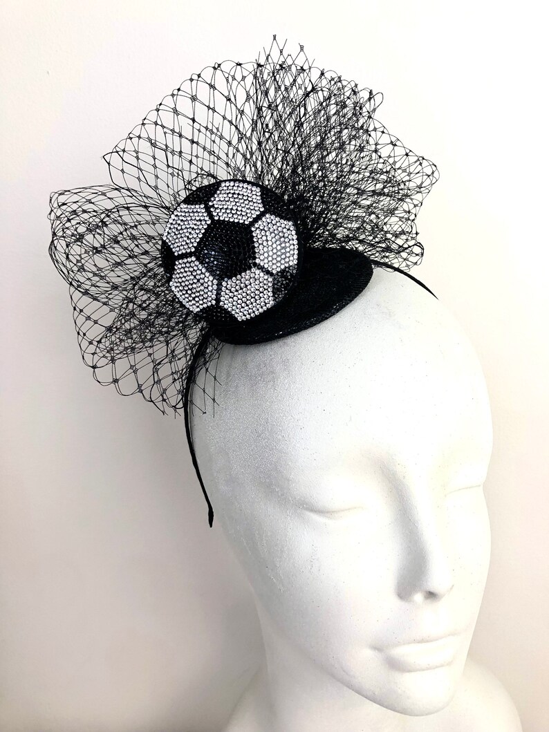 May include: A black headband with a decorative soccer ball design. The soccer ball is embellished with sparkling white and black rhinestones. A black netting is attached to the headband, creating a dramatic effect.