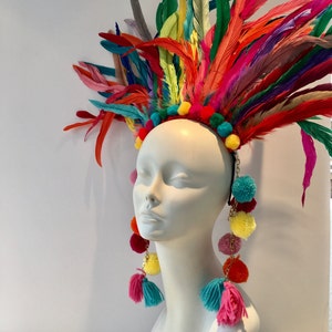 May include: A colorful feather headdress with pom poms and matching earrings. The headdress is made of a variety of feathers in shades of red, orange, yellow, green, blue, and purple. The earrings are made of pom poms in the same colors as the headdress.