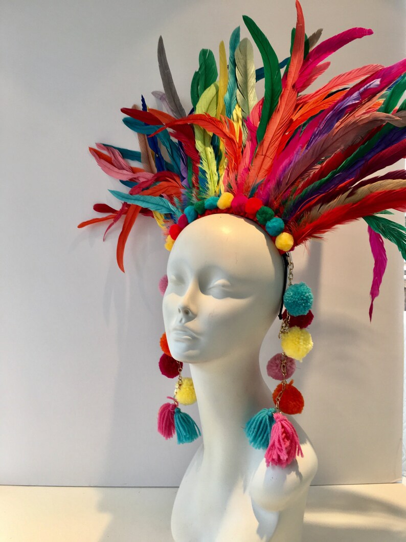 May include: A colorful feather headdress with pom poms and matching earrings. The headdress is made of a variety of feathers in shades of red, orange, yellow, green, blue, and purple. The earrings are made of pom poms in the same colors as the headdress.