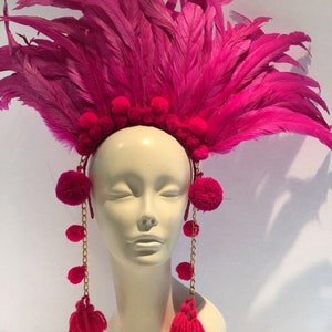 May include: A pink feather headdress with pom poms and gold chains. The headdress is worn on a white mannequin head.