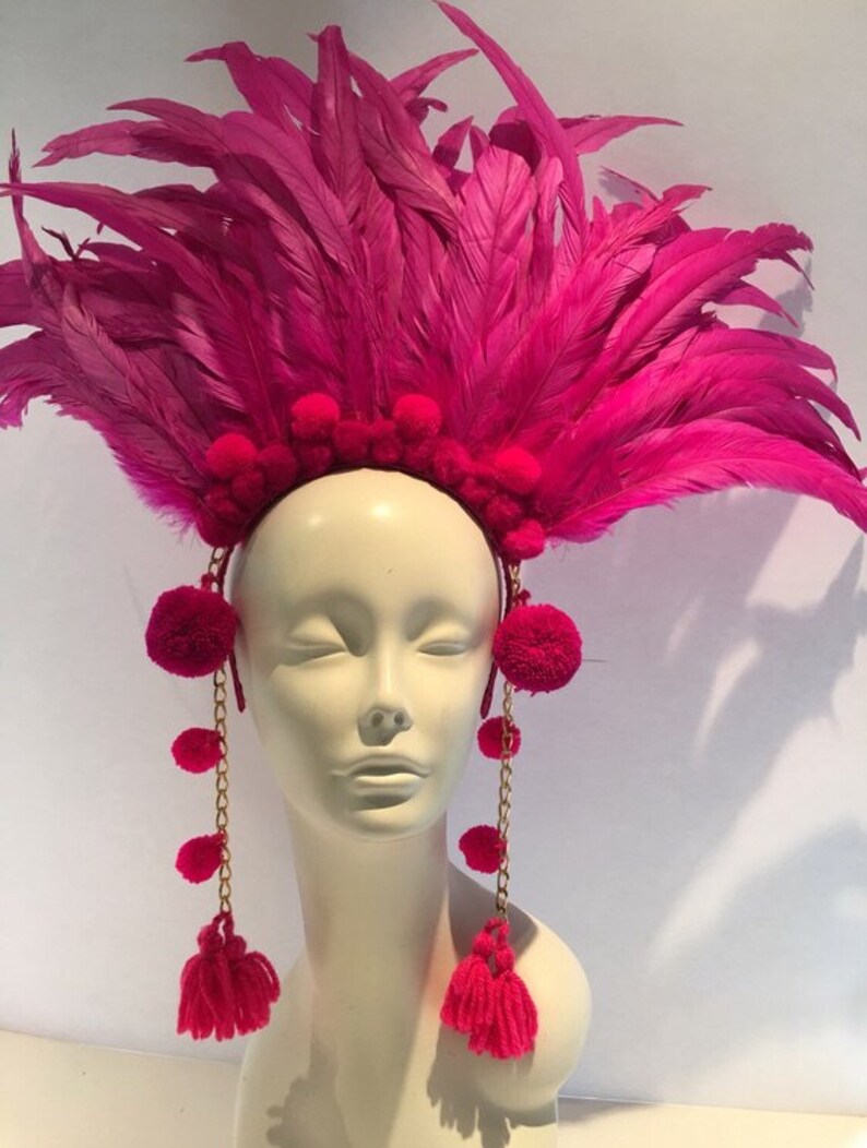 May include: A pink feather headdress with pom poms and gold chains. The headdress is worn on a white mannequin head.