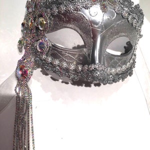 May include: A silver masquerade mask with a beaded fringe and iridescent rhinestones. The mask has a delicate, ornate design and is perfect for a costume party or special occasion.
