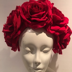 May include: A headpiece featuring a dense arrangement of large, deep red fabric roses. The flowers are clustered together to form a crown-like shape, covering the top and sides of a smooth, off-white, featureless mannequin head bust.