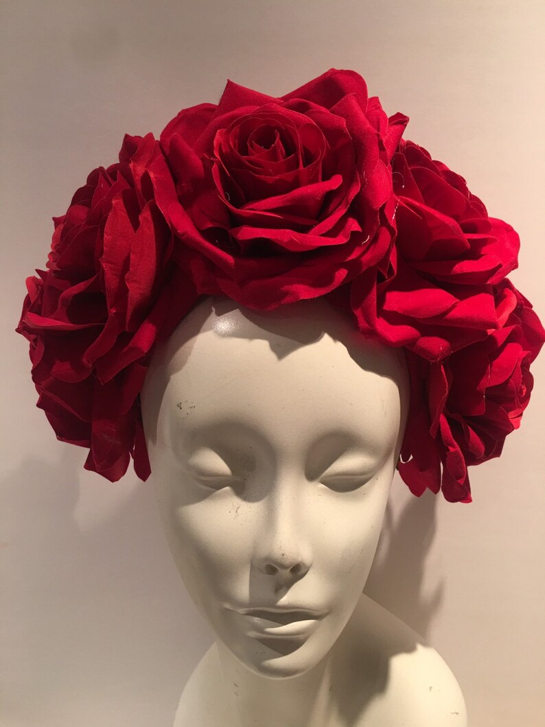 May include: A headpiece featuring a dense arrangement of large, deep red fabric roses. The flowers are clustered together to form a crown-like shape, covering the top and sides of a smooth, off-white, featureless mannequin head bust.