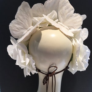 May include: White flower crown with a brown leather tie. The crown is made of paper flowers and is designed to be worn on the head.