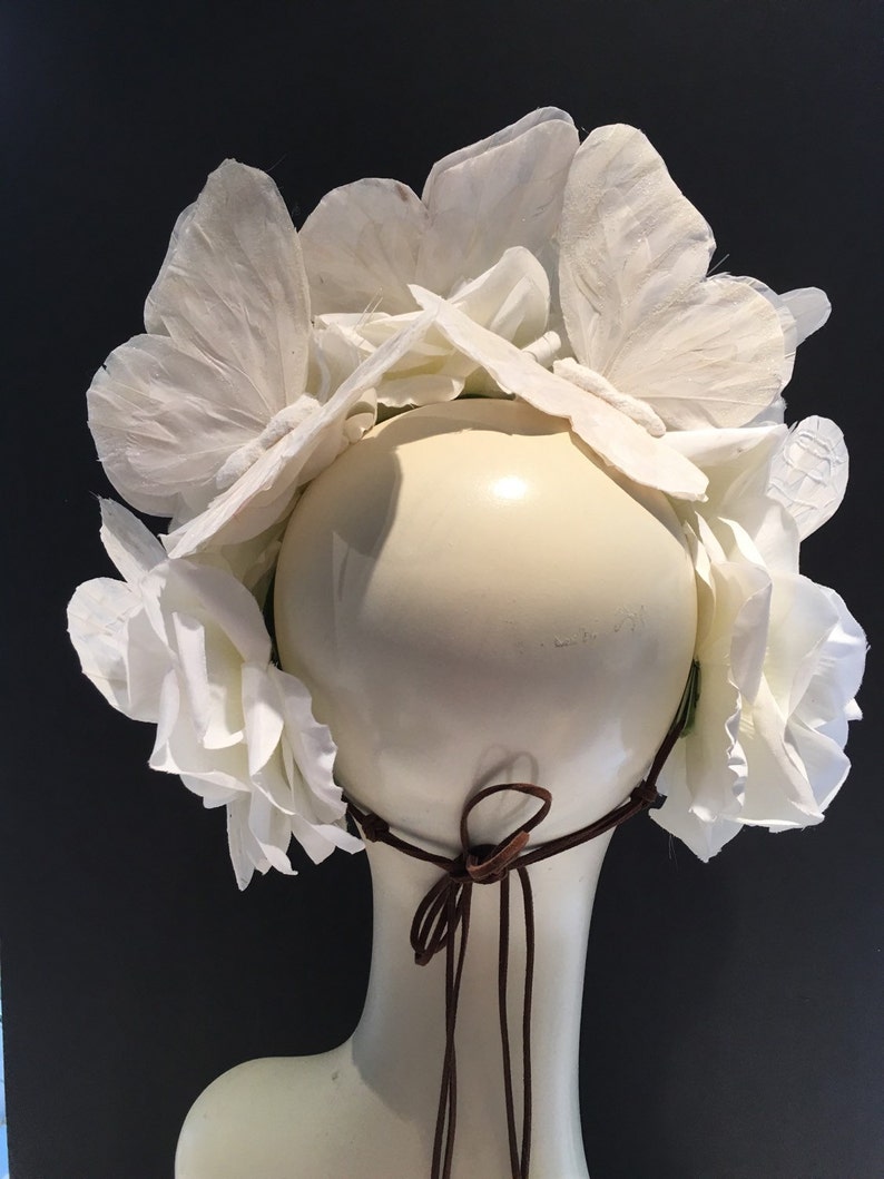 May include: White flower crown with a brown leather tie. The crown is made of paper flowers and is designed to be worn on the head.