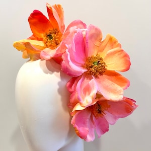 Puede incluir: Un tocado con tres grandes flores artificiales superpuestas en tonos vibrantes de rosa, naranja y amarillo. Los pétalos tienen una suave transición de color degradado y cada flor muestra un estambre central texturizado de color dorado.