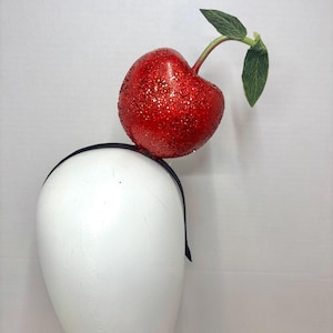 May include: A headband featuring a large, round, red glitter-covered cherry ornament. The cherry has a green stem and two green fabric leaves attached to the top. The headband itself is a thin, dark-colored band that sits securely.