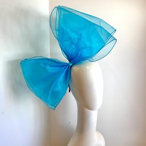 May include: A large, bright turquoise blue bow made of tulle fabric. The bow is worn on a mannequin's head as a hair accessory.