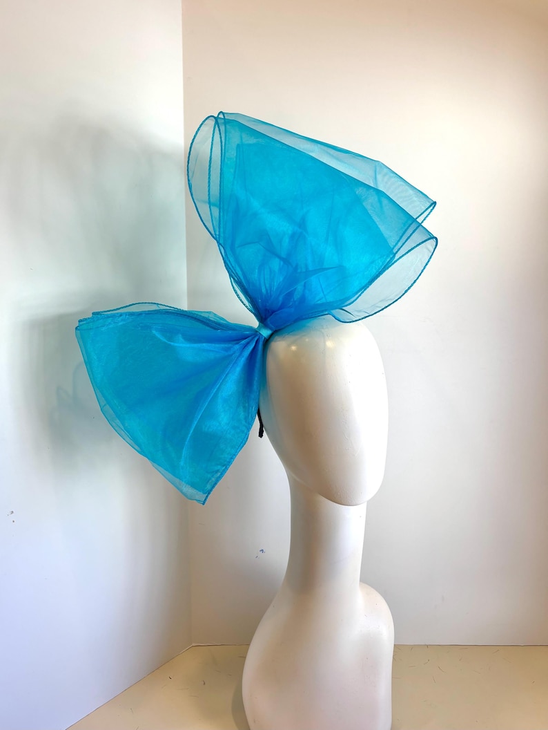 May include: A large, bright turquoise blue bow made of tulle fabric. The bow is worn on a mannequin's head as a hair accessory.