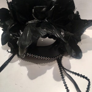 Puede incluir: Una m&aacute;scara negra de carnaval con un dise&ntilde;o floral de plumas. La m&aacute;scara est&aacute; adornada con peque&ntilde;os adornos brillantes a lo largo del borde inferior y presenta detalles colgantes de cuentas. El dise&ntilde;o se encuentra sobre un fondo blanco.