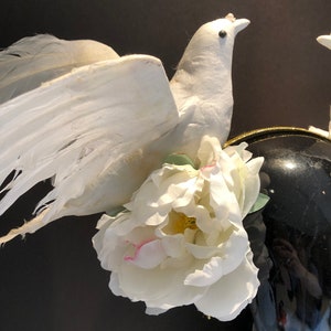 May include: A white decorative dove with outstretched wings, adorned with a large white peony flower. The dove is perched near a black, glossy vase, creating a contrast of textures and colors. The overall theme suggests a wedding or celebratory decor.