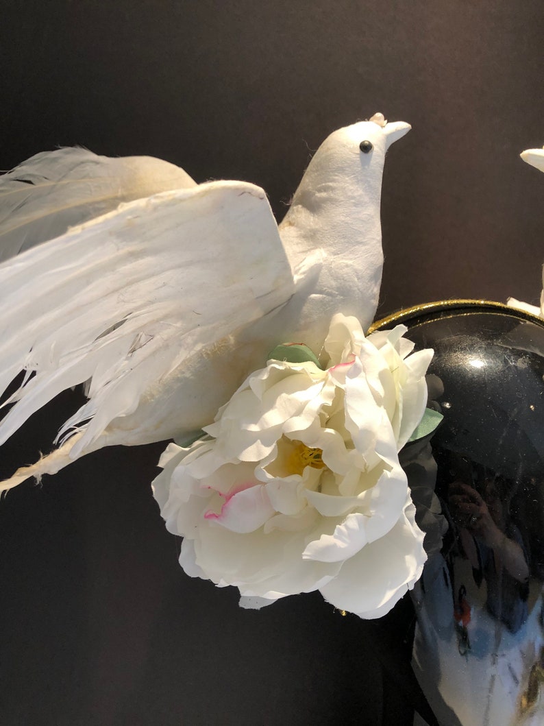 May include: A white decorative dove with outstretched wings, adorned with a large white peony flower. The dove is perched near a black, glossy vase, creating a contrast of textures and colors. The overall theme suggests a wedding or celebratory decor.