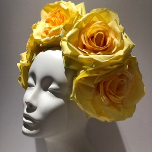 May include: A floral headpiece featuring multiple large, pale yellow artificial roses arranged in a cluster. The flowers are attached to a base that sits atop a smooth, white, featureless mannequin head, showcasing the layered petals of the blooms.