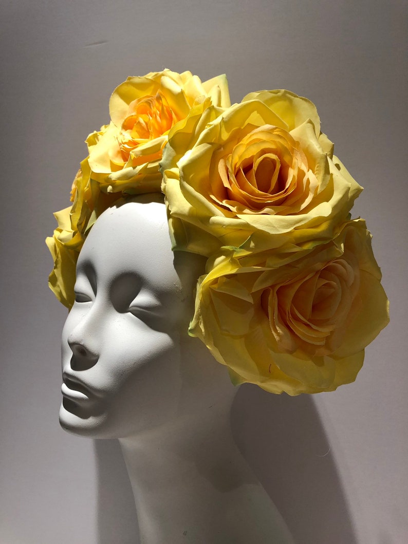 May include: A floral headpiece featuring multiple large, pale yellow artificial roses arranged in a cluster. The flowers are attached to a base that sits atop a smooth, white, featureless mannequin head, showcasing the layered petals of the blooms.
