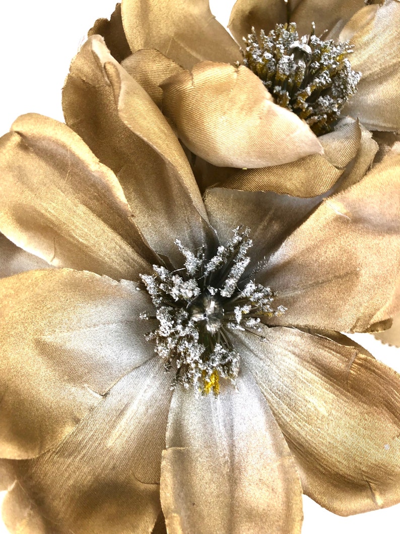K&ouml;nnte beinhalten: Nahaufnahme von zwei gro&szlig;en, k&uuml;nstlichen Blumen mit geschichteten, goldbraunen Bl&uuml;tenbl&auml;ttern. Die Zentren weisen eine Ansammlung von silberfarbenen, strukturierten Akzenten auf. Die Blumen dienen wahrscheinlich dekorativen Zwecken, m&ouml;glicherweise f&uuml;r die Heimdekoration oder zum Basteln.