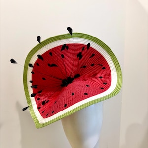 Puede incluir: Un sombrero caprichoso con forma de rodaja de sandía. El sombrero presenta un interior rojo con detalles de semillas negras, una corteza blanca y un borde exterior verde. Plumas negras están unidas al sombrero. El sombrero se exhibe en una cabeza de maniquí.