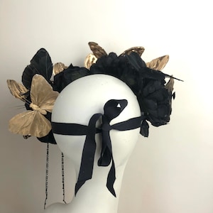 Puede incluir: Tocado decorativo con flores de tela negra, detalles de mariposas doradas y una cinta negra. El tocado se exhibe en una cabeza de maniqu&iacute; blanca, mostrando su dise&ntilde;o y detalles.
