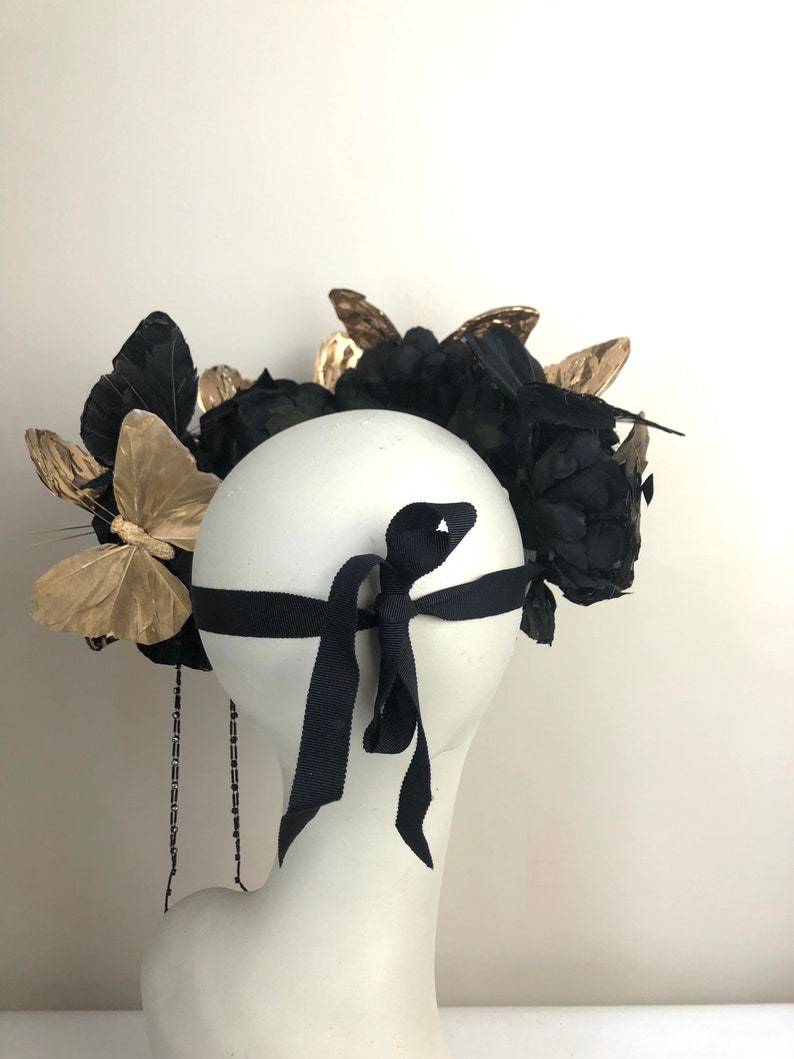 Puede incluir: Tocado decorativo con flores de tela negra, detalles de mariposas doradas y una cinta negra. El tocado se exhibe en una cabeza de maniqu&iacute; blanca, mostrando su dise&ntilde;o y detalles.