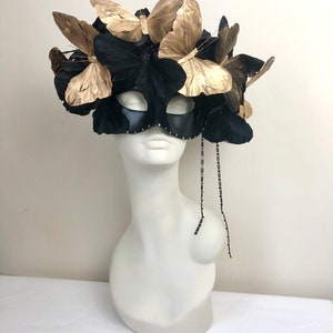 Puede incluir: Una m&aacute;scara negra adornada con mariposas doradas y plumas negras. La m&aacute;scara tiene un fleco de cuentas y se exhibe en una cabeza de maniqu&iacute; blanca. Un accesorio decorativo.