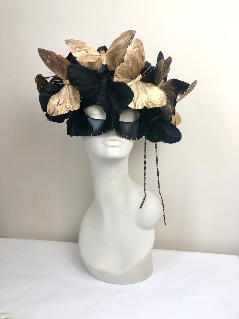 Puede incluir: Una m&aacute;scara negra adornada con mariposas doradas y plumas negras. La m&aacute;scara tiene un fleco de cuentas y se exhibe en una cabeza de maniqu&iacute; blanca. Un accesorio decorativo.