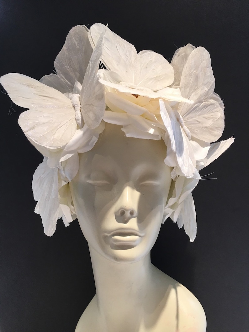 May include: A white headpiece made of paper butterflies. The butterflies are arranged in a circular pattern around the head. The headpiece is worn on a white mannequin head.