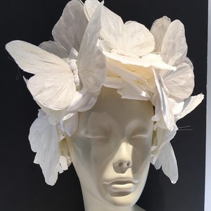 May include: A white headpiece made of paper butterflies. The butterflies are arranged in a circular pattern around the head. The headpiece is worn on a white mannequin head.