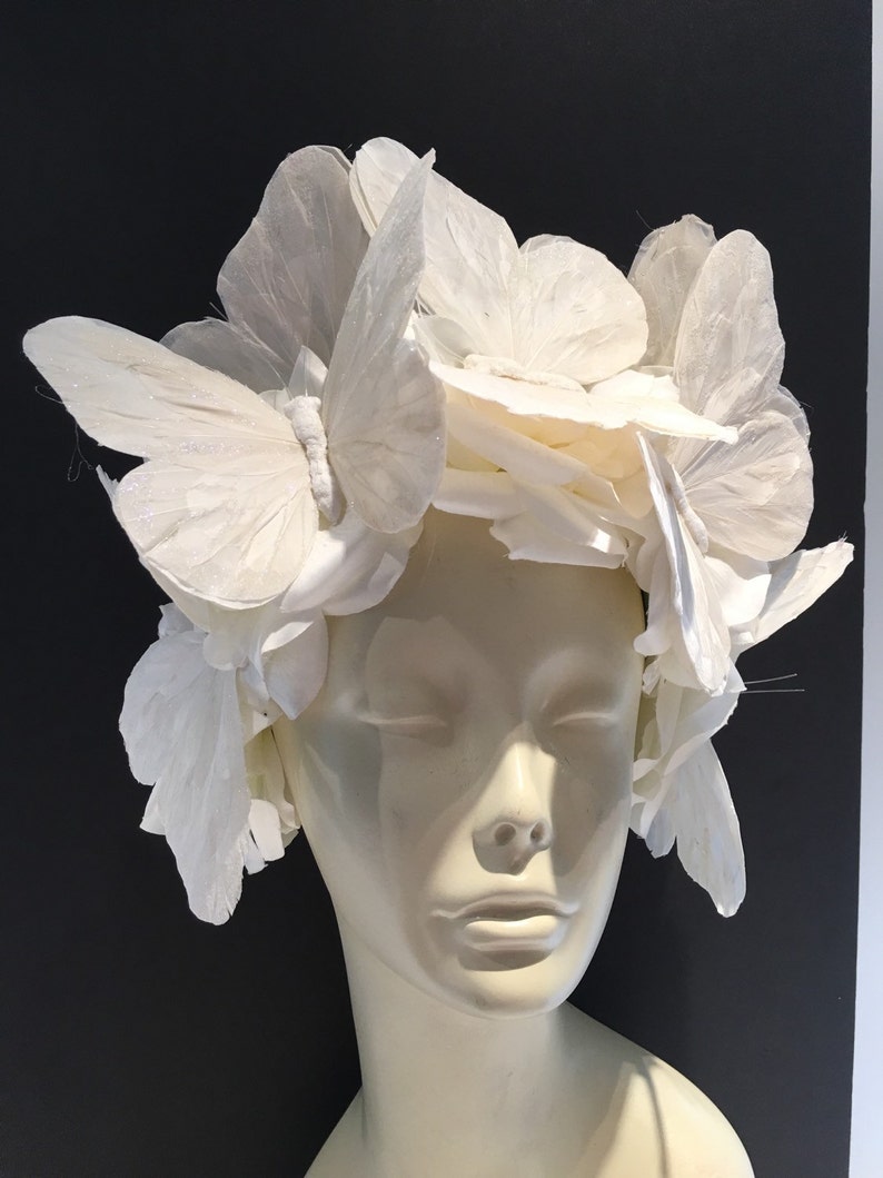 May include: A white headpiece made of paper butterflies. The butterflies are arranged in a circular pattern around the head. The headpiece is worn on a white mannequin head.