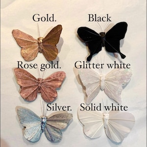 Puede incluir: Adornos decorativos de mariposas en oro, negro, oro rosa, blanco brillante, plata y blanco liso. Cada mariposa tiene alas detalladas y un alambre para colgar. Las etiquetas indican los colores.