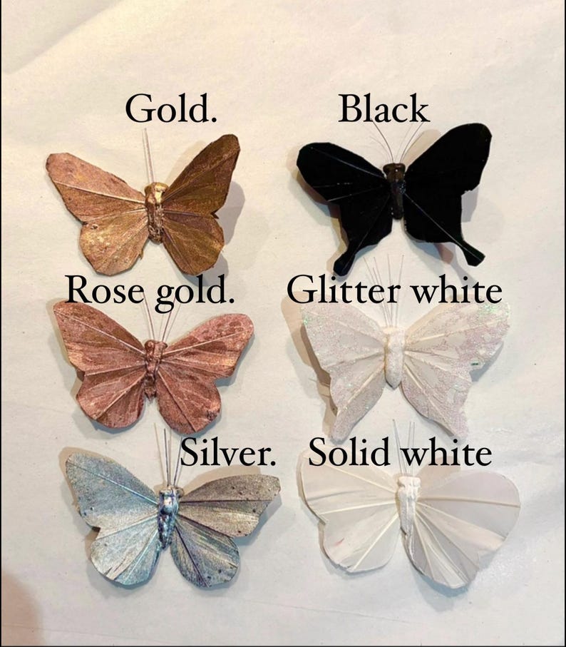 Puede incluir: Adornos decorativos de mariposas en oro, negro, oro rosa, blanco brillante, plata y blanco liso. Cada mariposa tiene alas detalladas y un alambre para colgar. Las etiquetas indican los colores.