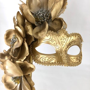 K&ouml;nnte beinhalten: Eine goldfarbene Maske f&uuml;r einen Maskenball mit floralen Akzenten. Die Maske hat ein verziertes Design und ist an einem goldenen Griff befestigt, der mit gro&szlig;en, goldenen Stoffblumen mit silbernen Zentren verziert ist. Ideal f&uuml;r einen Maskenball.