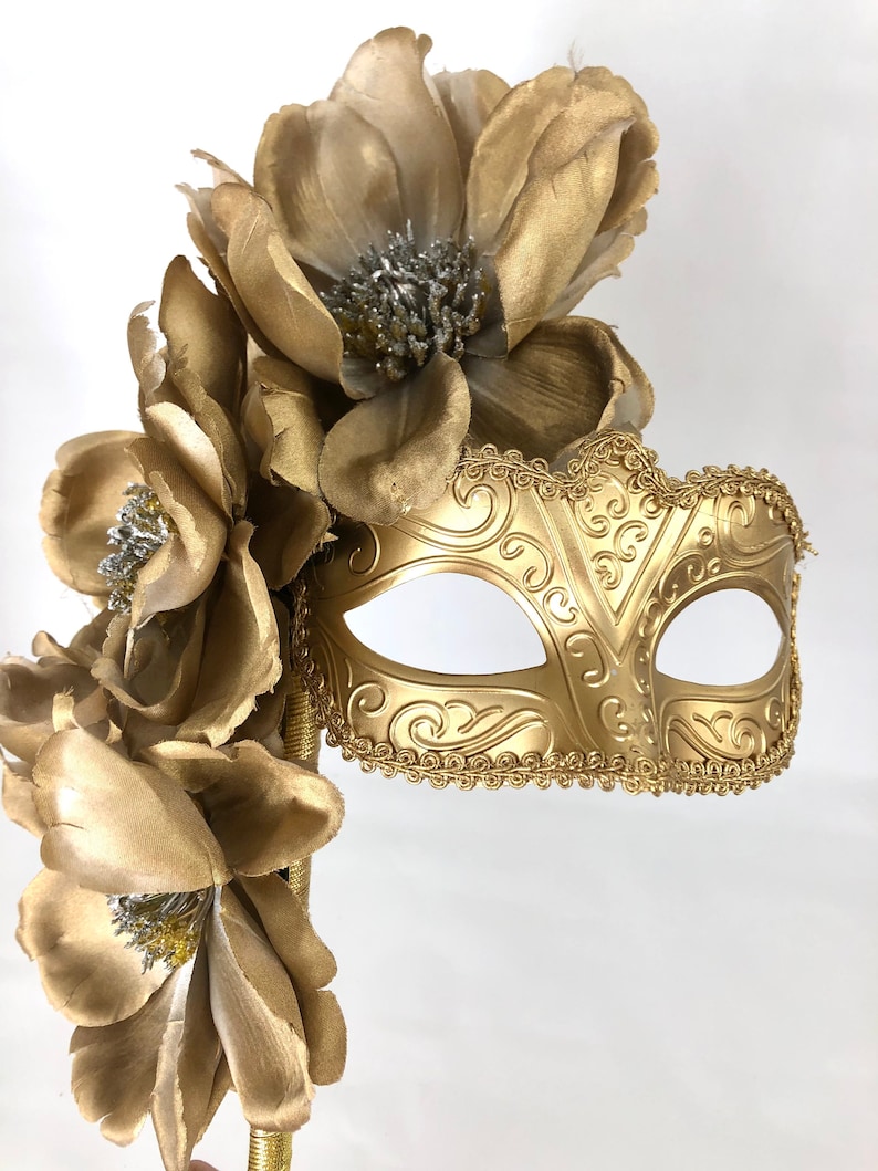 K&ouml;nnte beinhalten: Eine goldfarbene Maske f&uuml;r einen Maskenball mit floralen Akzenten. Die Maske hat ein verziertes Design und ist an einem goldenen Griff befestigt, der mit gro&szlig;en, goldenen Stoffblumen mit silbernen Zentren verziert ist. Ideal f&uuml;r einen Maskenball.