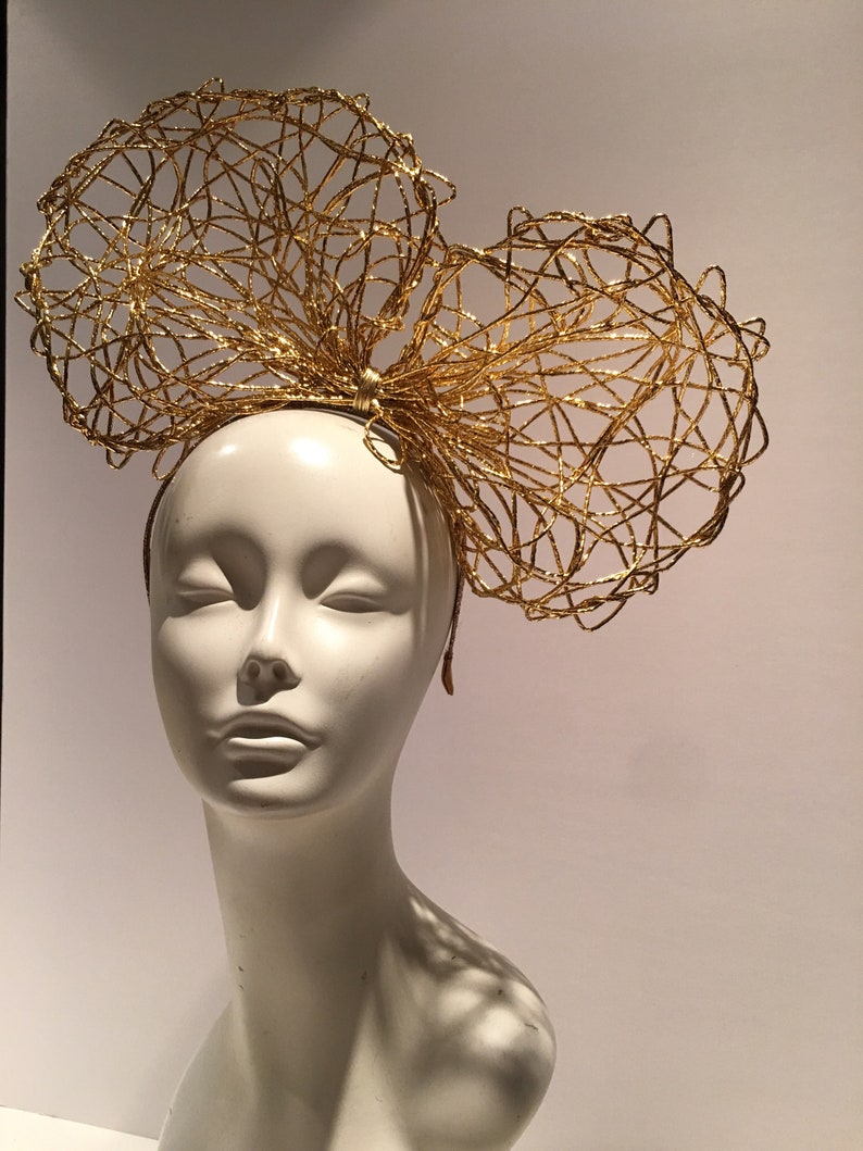 May include: A gold wire headband with a large bow on top. The bow is made of multiple layers of wire that are intertwined to create a delicate and intricate design.