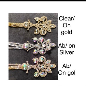 May include: Three rhinestone brooches with dangling chains. The brooches are in the shape of a floral design. The top brooch has clear rhinestones on a gold base. The middle brooch has iridescent rhinestones on a silver base. The bottom brooch has iridescent rhinestones on a gold base.