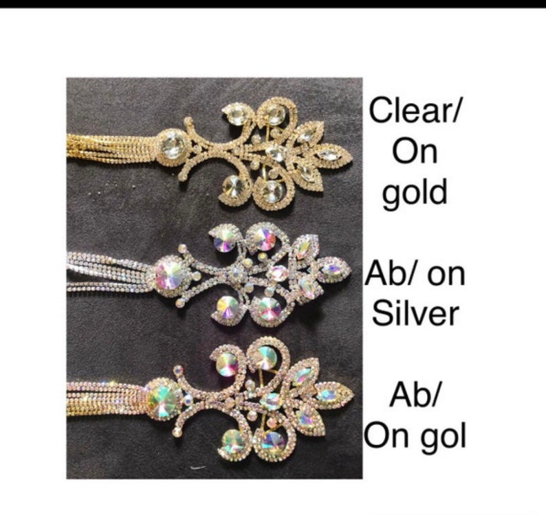 May include: Three rhinestone brooches with dangling chains. The brooches are in the shape of a floral design. The top brooch has clear rhinestones on a gold base. The middle brooch has iridescent rhinestones on a silver base. The bottom brooch has iridescent rhinestones on a gold base.