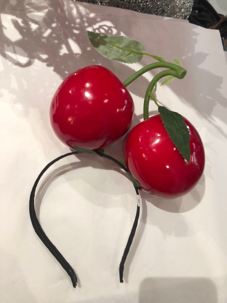 K&ouml;nnte beinhalten: Ein schwarzes Stirnband mit zwei gro&szlig;en, gl&auml;nzenden roten Kirschen. Die Kirschen haben gr&uuml;ne Stiele und Bl&auml;tter. Das Stirnband besteht aus einem d&uuml;nnen, flexiblen Material, die Kirschen aus gl&auml;nzendem Material.