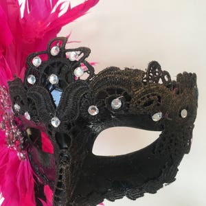 May include: A black masquerade mask with a pink feather accent. The mask is decorated with black lace and rhinestones.