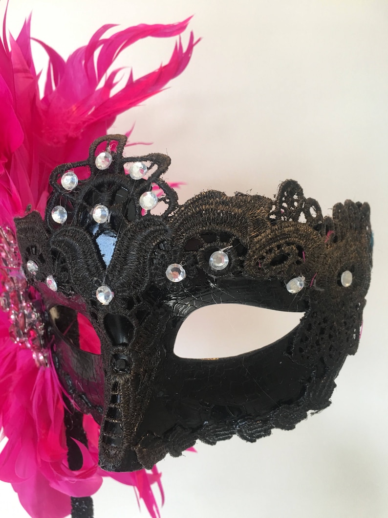 May include: A black masquerade mask with a pink feather accent. The mask is decorated with black lace and rhinestones.
