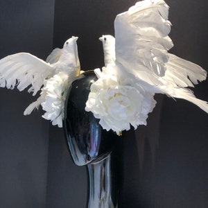 May include: A black mannequin head adorned with a white floral and dove-themed headpiece. The headpiece features two white doves with outstretched wings, surrounded by white flowers. The headpiece is set against a black background.