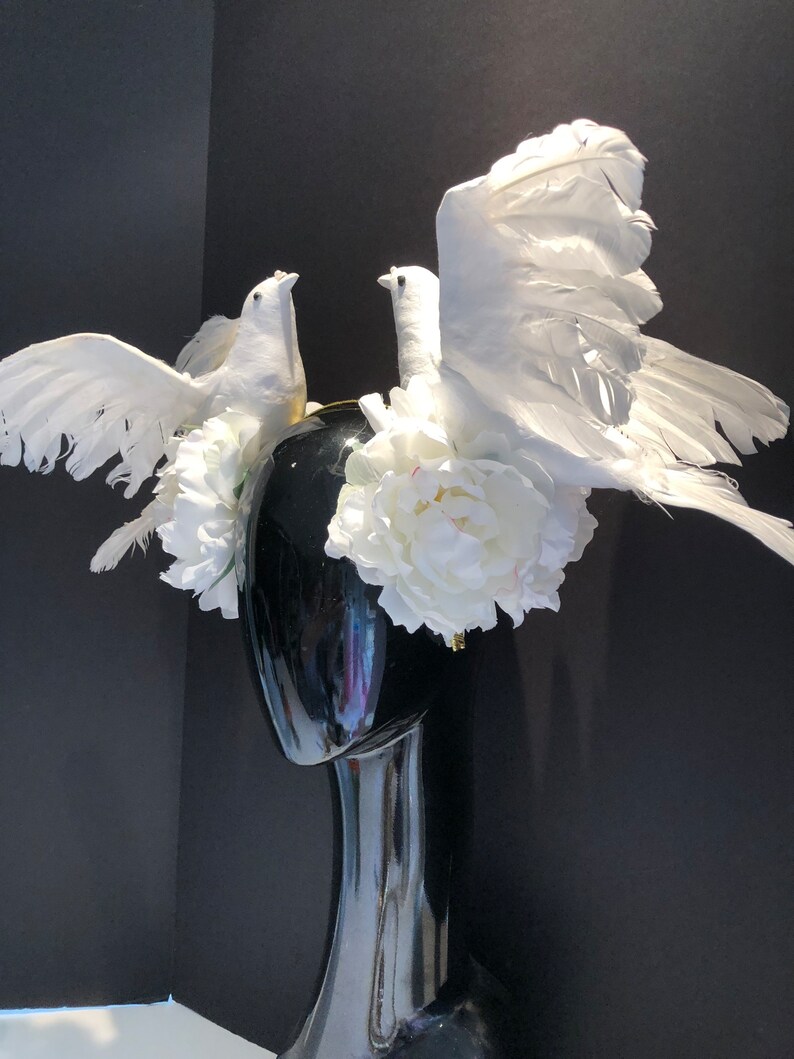 May include: A black mannequin head adorned with a white floral and dove-themed headpiece. The headpiece features two white doves with outstretched wings, surrounded by white flowers. The headpiece is set against a black background.