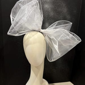 May include: A large white tulle bow headband. The bow is made of multiple layers of tulle and is tied in a large, dramatic knot. The headband is black.