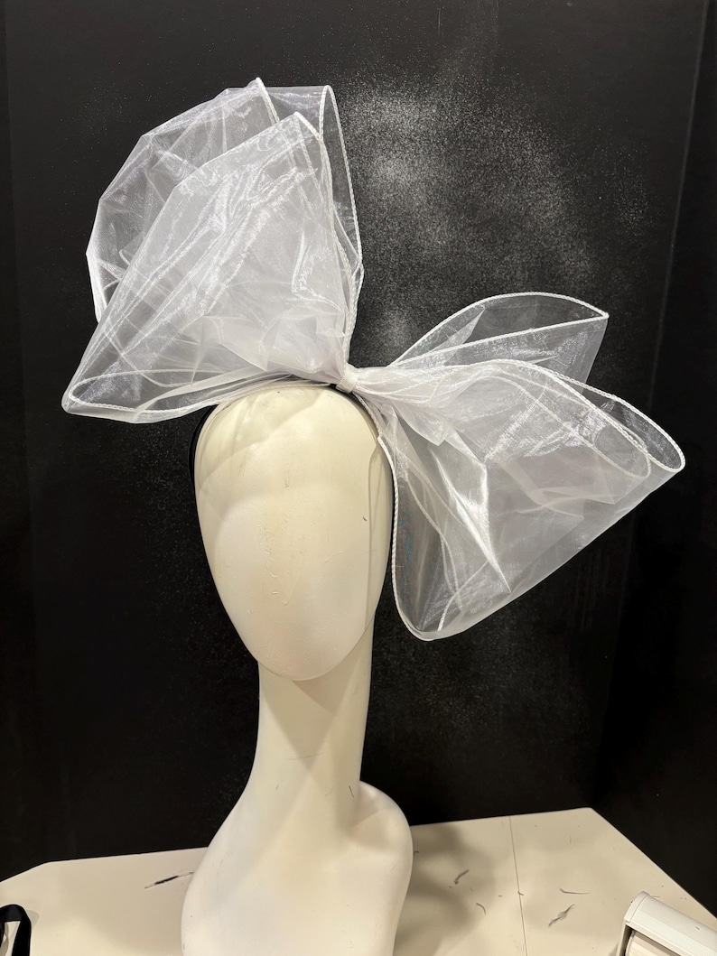 May include: A large white tulle bow headband. The bow is made of multiple layers of tulle and is tied in a large, dramatic knot. The headband is black.